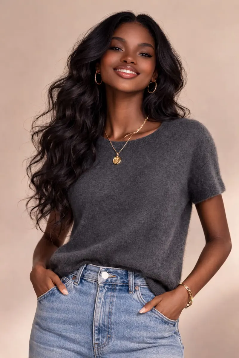 Smiling woman in a charcoal short-sleeve knit top with high-waisted light blue denim, gold hoop earrings and layered necklace, studio background.