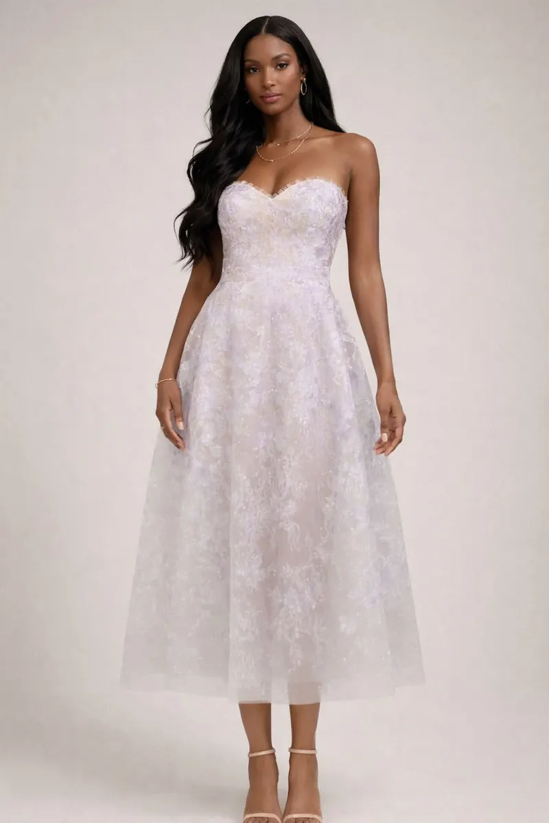 Full-body studio portrait of a Black woman with long wavy hair wearing a strapless lavender tulle midi dress with subtle floral texture and nude strappy heels on a beige background.