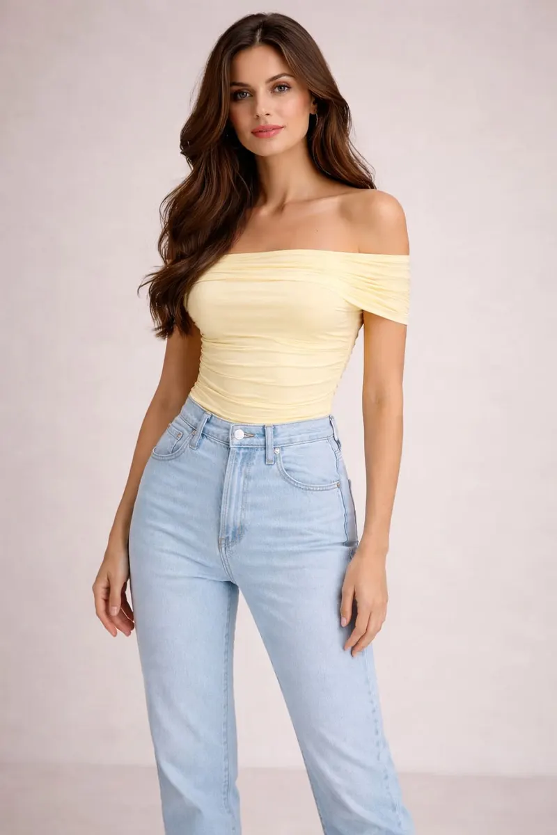 Brunette model wearing a pastel yellow off-shoulder ruched top with high-waisted light-wash straight-leg jeans in a neutral studio setting.
