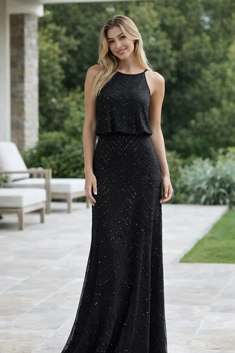 Full-length portrait of a woman wearing a sleeveless black embellished evening gown with a soft draped bodice and floor-length sheer overlay in an outdoor editorial setting