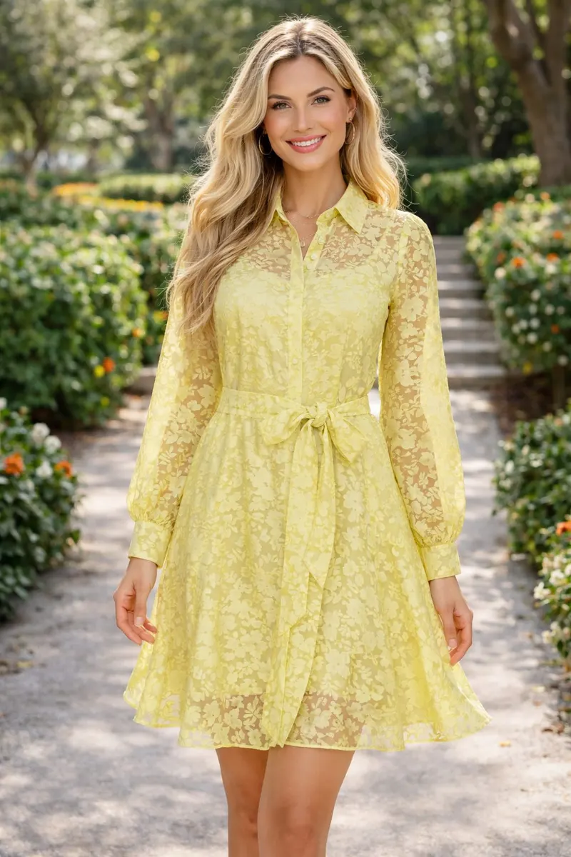 Blonde woman wearing a lemon-yellow sheer floral shirt dress with a waist tie and long sleeves, styled with layered gold jewelry in a garden walkway