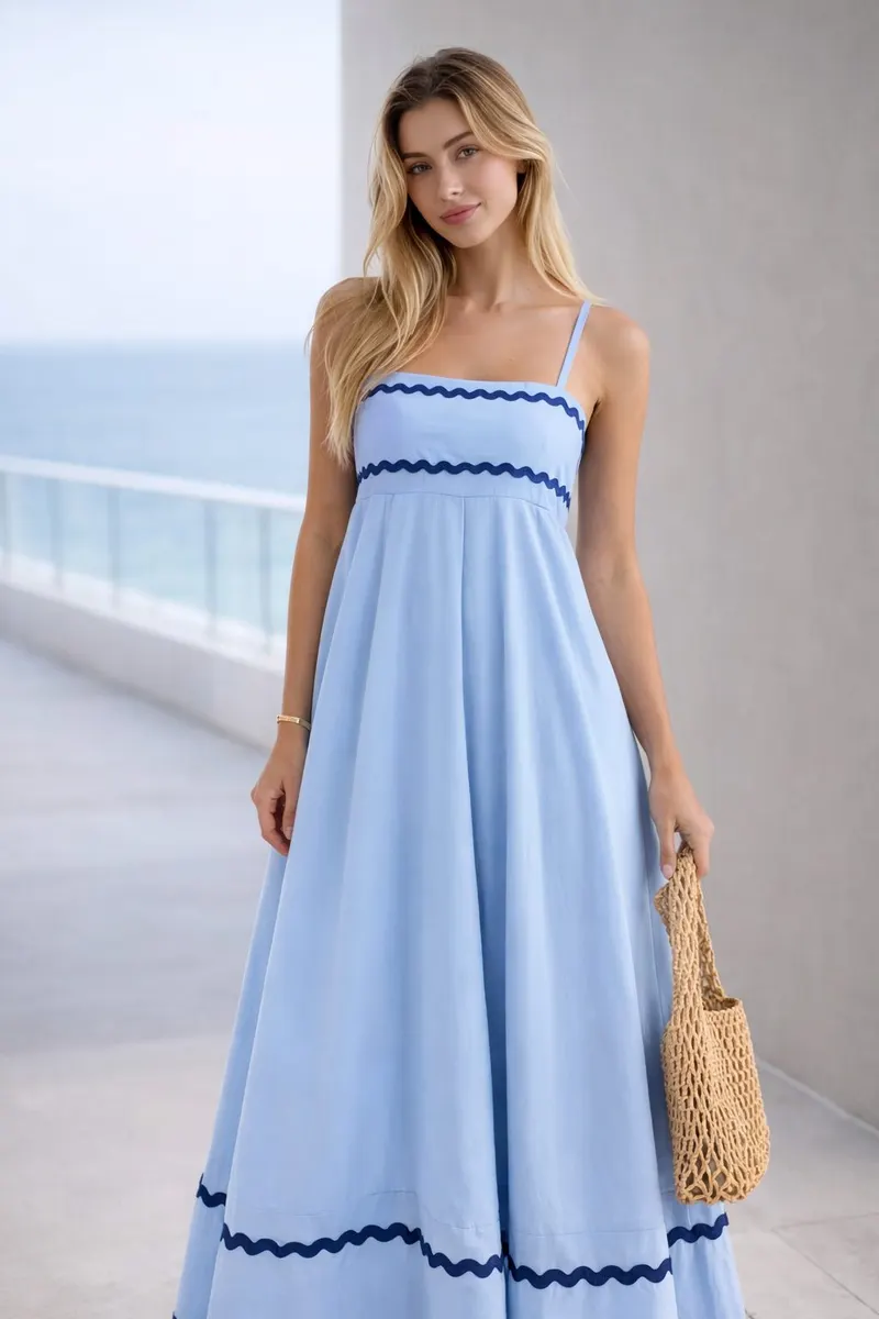 Woman wearing a light blue sleeveless maxi dress with contrasting wave trim and a woven bag in a modern editorial setting